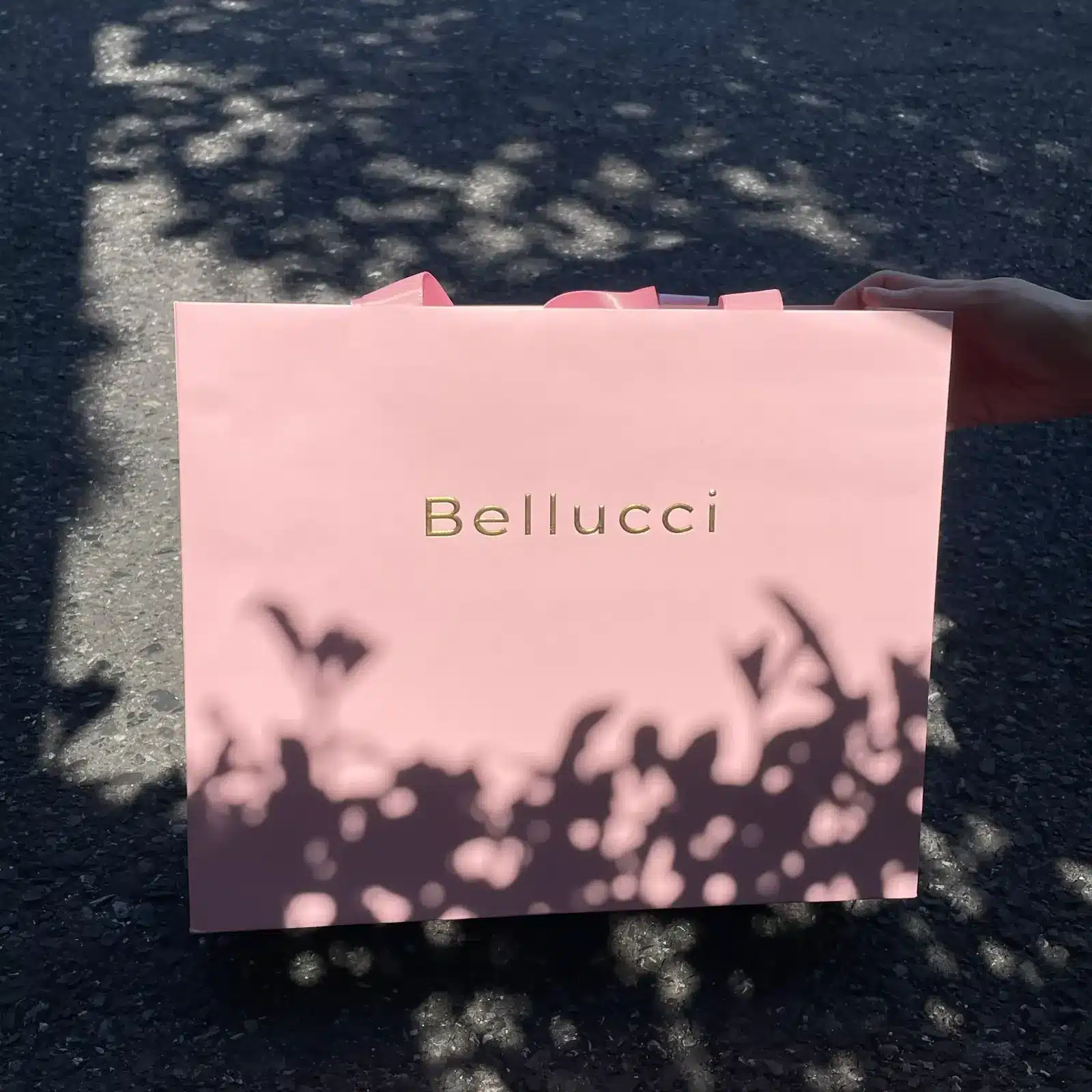 Front view of Bellucci pink shopping bag with gold‑foil logo under natural daylight