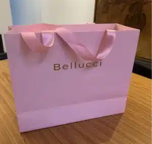 Real‑life photo of Bellucci blush‑pink luxury shopping bag with gold logo and satin ribbon handles on wood table